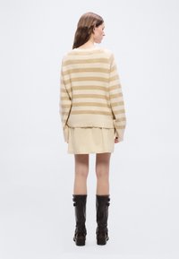 Γυναίκα γυρισμένη με την πλάτη φοράει μπεζ και κρεμ ριγέ πουλόβερ, κρεμ φούστα και μαύρες μπότες έως το γόνατο σε απλό λευκό φόντο.