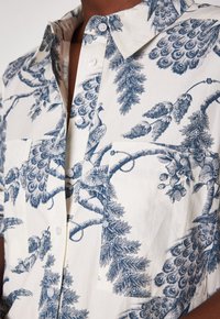 Camicia a maniche corte con bottoni, stampa di pavone blu e fiori su tessuto bianco, indossata da una persona con pelle scura.