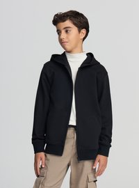 Felpa nera con zip realizzata in tessuto morbido, dotata di tasca a marsupio e cappuccio con coulisse, indossata sopra una maglietta bianca e pantaloni cargo beige.