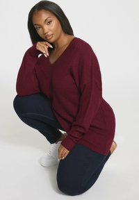 Bordowy sweter prążkowany z dekoltem w kształcie V, o luźnym kroju i długich rękawach, zestawiony z ciemnoniebieskimi dżinsami i białymi trampkami.