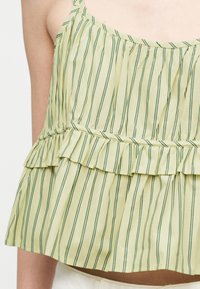 Haut jaune sans manches avec de fines rayures verticales vertes et un ourlet à volants porté par une personne cadrée au niveau du torse.