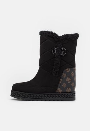 Botte d’hiver matelassée noire avec bordure en shearling, détail chaîne sur la semelle et panneau de talon marron à motifs avec le logo de la marque.