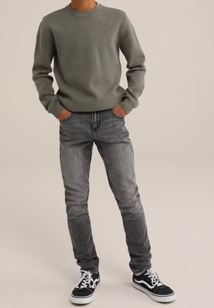 Personne portant un pull côtelé vert, un jean noir délavé et des baskets noires et blanches à lacets, debout les mains dans les poches.