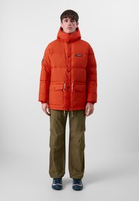Pufferjakke i lys orange med høy krage, glidelås foran, to frontlommer og hem med snøring. Kombinert med khaki bukser og blå joggesko.