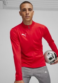 Rode sportjack met gestructureerde stof en ritssluiting bij de hals, voorzien van een klein wit logo. Het model houdt een zilveren voetbalsbal vast.