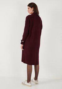 Femeie îmbrăcată cu o rochie lungă din tricot rib din burgund, colanți negri transparenți, adidași albi și cercei rotunzi din argint, stând în fața unui fundal simplu.