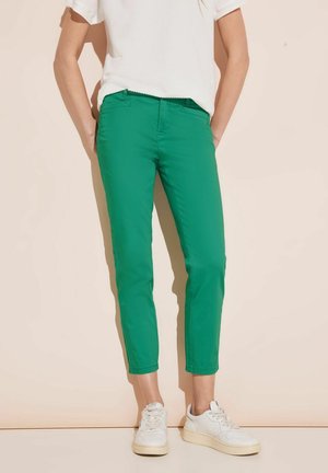 Pantalons en mélange de coton vert avec une coupe slim, longueur cropped et poches latérales, associés à des baskets blanches sur un fond clair.