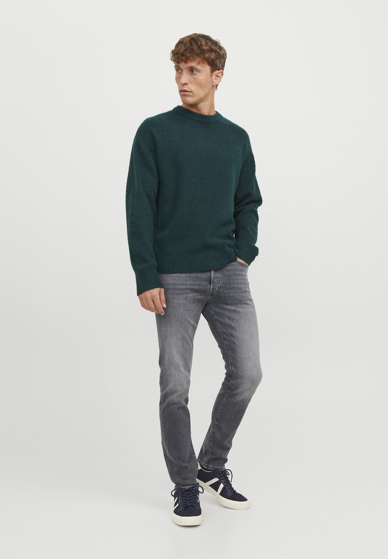 Jersey de punto verde oscuro con cuello redondo, tejido texturizado, puños y dobladillo acanalados, combinado con unos pantalones gris ajustados y zapatillas marinas.