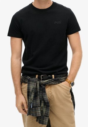 Mand iført en sort t-shirt med korte ærmer, beige bukser, en ternet skjorte bundet om livet og et armbåndsur på venstre håndled.