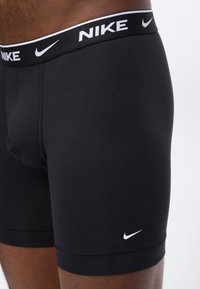 Μαύρα boxer briefs της Nike με λευκό λογότυπο swoosh στον αριστερό μηρό και λογότυπο Nike στη μέση, φορεμένα από ένα άτομο.