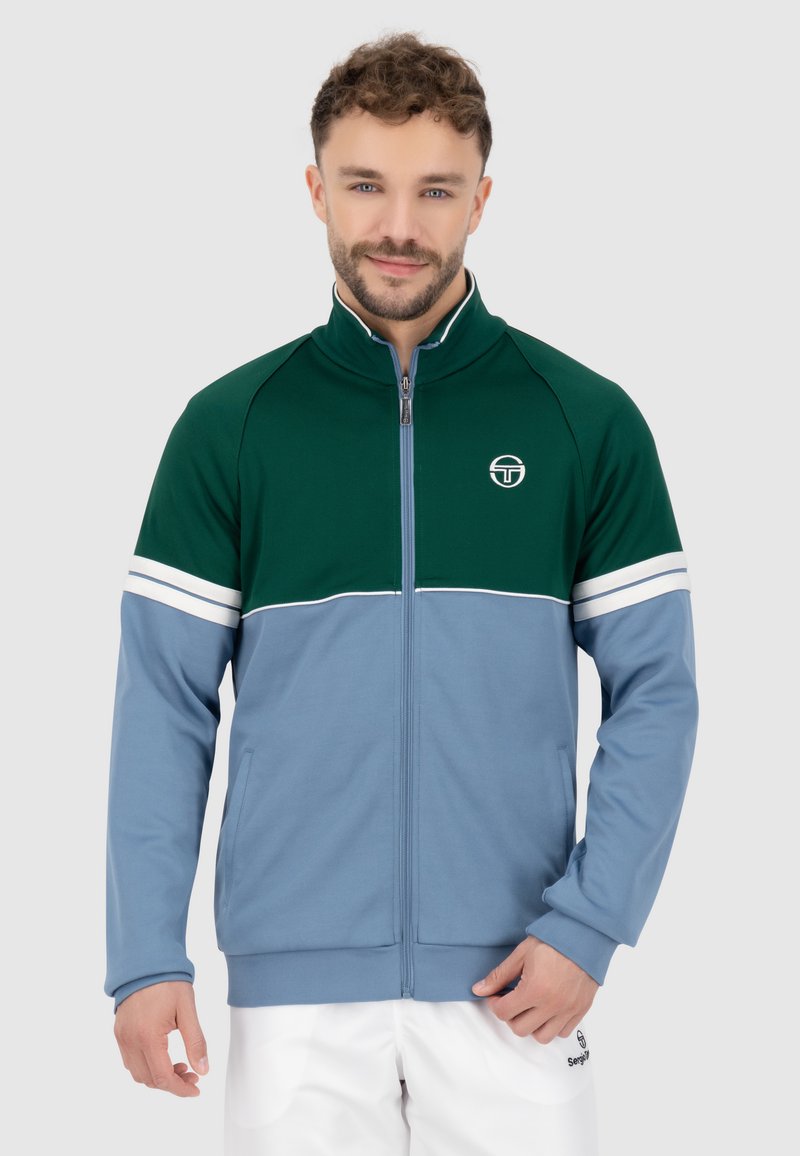 Giacca a track con zip in verde scuro e azzurro; presenta strisce bianche sulle maniche, colletto alto e logo sul petto. Tessuto morbido.