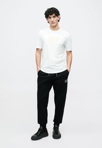 Tricou alb cu mânecă scurtă și logo auriu, asortat cu pantaloni negri lejeri. Design minimalist, textură netedă și adidași negri.