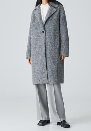 Personne portant un long manteau gris texturé sur un haut blanc, un pantalon large gris clair et des chaussures noires sur un fond clair uni.