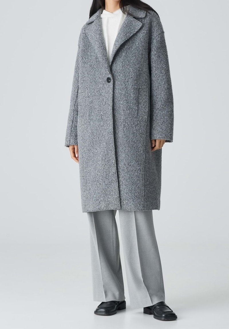 Personne portant un long manteau gris texturé sur un haut blanc, un pantalon large gris clair et des chaussures noires sur un fond clair uni.