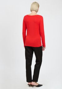 Rotes Langarmoberteil mit weichem Material und runder Ausschnittlinie, kombiniert mit schwarzen Hosen und schwarzen spitzen High Heels.