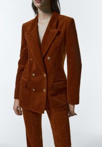 Blazer croisé en velours côtelé couleur rouille avec boutons dorés et revers crantés, porté avec un pantalon en velours côtelé assorti. Surface texturée visible.