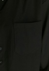 Tissu de chemise noire avec une poche poitrine visible et deux boutons sur le côté gauche de l’image.