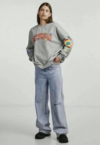 Γκρι φούτερ με την επιγραφή «ΣΑΒΒΑΤΟ» σε πολύχρωμα γράμματα, διακοσμημένο με σχέδια χαρούμενων προσώπων και λουλουδιών, σε συνδυασμό με ανοιχτού μπλε φαρδιά τζιν και μαύρες παπούτσια.