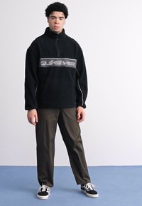 Crni flis pulover s patentnim zatvaračem na vratu, sivi Quiksilver logo na prsima; uparen s tamnosmešnim hlačama i crnim tenisicama.