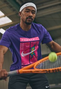 Άνδρας που φοράει ένα μωβ μπλουζάκι Nike και λευκή κορδέλα, χτυπάει μια μπάλα τένις με μια πορτοκαλί ρακέτα σε κλειστό χώρο.