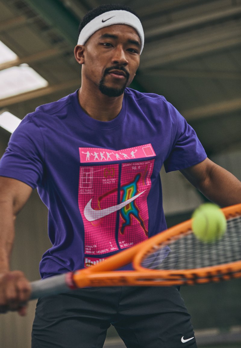 Άνδρας που φοράει ένα μωβ μπλουζάκι Nike και λευκή κορδέλα, χτυπάει μια μπάλα τένις με μια πορτοκαλί ρακέτα σε κλειστό χώρο.