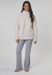 Ljust rosa fleece-pullover med hög krage och framficka, parat med grå utsvängda byxor. Texturerad tyg och sidofickor är synliga.