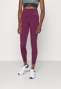Kobieta ubrana w purpurowe legginsy Nike o wysokim stanie z bocznymi kieszeniami, szary crop top i białe sneakersy Nike z granatowymi akcentami, stojąca na białej podłodze.