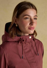 Bordowa kurtka z wysokim kołnierzem, z regulowanymi sznurkami, napami i gładką fakturą. Widoczne subtelne detale przeszycia.