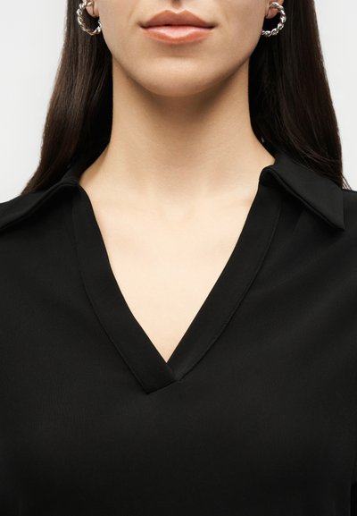 Γυναίκα που φοράει μαύρο τοπ με V λαιμόκοψη και ασημένια στριφογυριστά σκουλαρίκια κρίκους, με ίσια σκούρα μαλλιά ορατά πίσω από τους ώμους.