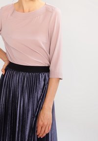 Donna che indossa una blusa rosa chiaro a tre quarti, infilata in una gonna plissettata viola scuro lucida con cintura nera.