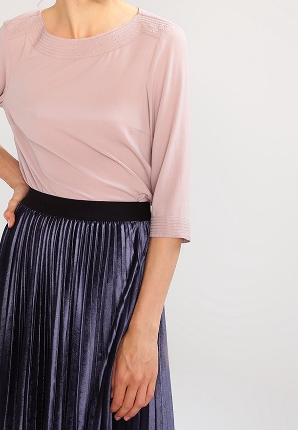 Donna che indossa una blusa rosa chiaro a tre quarti, infilata in una gonna plissettata viola scuro lucida con cintura nera.