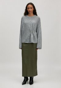 Grijze, oversized gebreide trui met een ronde hals en twee metalen accenten aan de voorkant, gecombineerd met een getextureerde olijfgroene midi-rok en zwarte enkellaarsjes.
