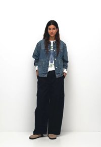 Jachetă din denim cu închidere prin butoane, purtată peste un hanorac alb cu grafică, asociată cu pantaloni cargo largi negri și pantofi slip-on maro.