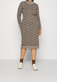 Robe en tricot marron avec un motif géométrique contrastant. Manches longues, col rond, poignets et ourlet côtelés. Associée à des chaussures à plateforme blanches.