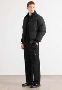 Black puffer jacket with a high collar, zip closure, and side pockets. Paired with black cargo pants featuring buttoned side pockets and black shoes.