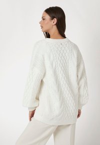 Witte gebreide trui met een kabelpatroon, oversized silhouette en geribde boorden. De achterkant heeft een klein gouden accent bij de hals.