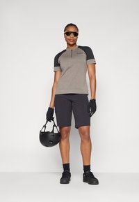 Grå kortermet skjorte med svarte detaljer og glidelås, sammen med svarte shorts. Modellen har på seg hansker og holder en svart hjelm. Solbriller inkludert.