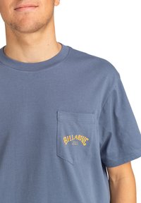 Modrá bavlněná tričko s kulatým výstřihem, krátkými rukávy a náprsní kapsou, na které je žlutý nápis a logo "Billabong". Hladká textura.