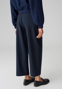 Pantalons larges bleu marine en tissu doux, dotés de deux poches arrière. Stylés avec une coupe décontractée et portés avec des chaussures noires à enfiler.