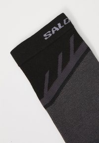 Calcetín deportivo negro y gris oscuro con un patrón de zigzag y una parte de texto cerca del borde superior sobre un fondo blanco liso.