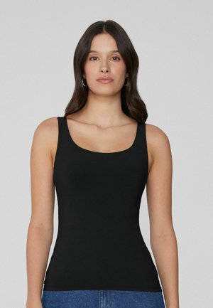 Vrouw met lang donker haar die zilveren oorringen draagt, een zwarte mouwloze top en blauwe spijkerbroek, kijkend naar voren tegen een effen grijze achtergrond.