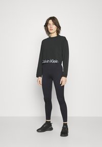 Felpa corta nera con il logo "Calvin Klein" all'orlo, abbinata a leggings neri aderenti e scarpe sportive nere. Tessuto morbido, design attillato.