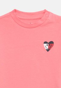 Ροζ μπλούζα με κοντά μανίκια, στρογγυλή λαιμόκοψη και ένα μικρό ασπρόμαυρο γραφικό καρδιά με χαμογελαστά  πρόσωπα στο αριστερό στήθος.