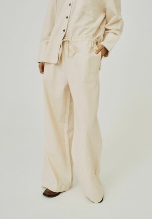 Pantaloni in lino beige, leggeri, con cintura elasticizzata, cordoncino e gambe larghe. Indossati con una camicia abbinata e scarpe marroni.