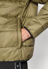 Giacca puffer verde oliva, caratterizzata da sezioni trapuntate, tessuto testurizzato e polsini neri. La mano sinistra è appoggiata nella tasca laterale.