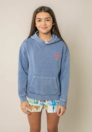 Fille aux longs cheveux foncés portant un sweat à capuche bleu délavé avec poche et un short coloré à motifs, debout devant un fond beige.