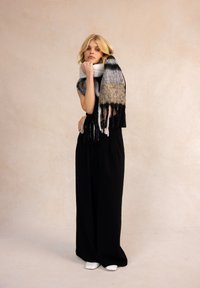 Écharpe en laine multicolore avec des rayures noires, blanches et grises ; présente des bords à franges. Le modèle porte une tenue noire et des chaussures blanches.