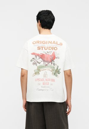 Man wearing white T-shirt with red cardinal bird and text "ORIGINALS STUDIO, Upstate New York 10553," standing facing away.
