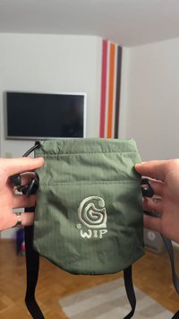 Žalia audinio rankinė su tekstūruotu raštu, reguliuojama juoda petneša ir siuvinėta logotipu priekyje. Viršuje raišteliu užsegama.