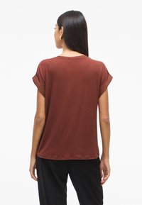 Top a maniche corte color ruggine con una vestibilità rilassata e maniche risvoltate, realizzato in un tessuto liscio. La vista posteriore mostra un design semplice e pulito.